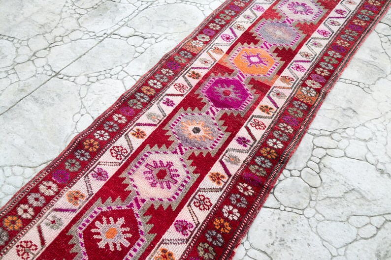May include: A long, narrow red and white runner rug with a geometric pattern in shades of pink, orange, and gray. The rug has a traditional Turkish design.
