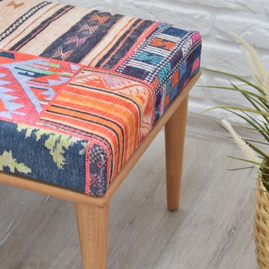 Pode incluir: Banqueta de madeira com assento em patchwork. O assento apresenta v&aacute;rios padr&otilde;es de tecido coloridos, incluindo motivos geom&eacute;tricos e florais. A banqueta tem quatro pernas de madeira clara e est&aacute; perto de uma planta decorativa.