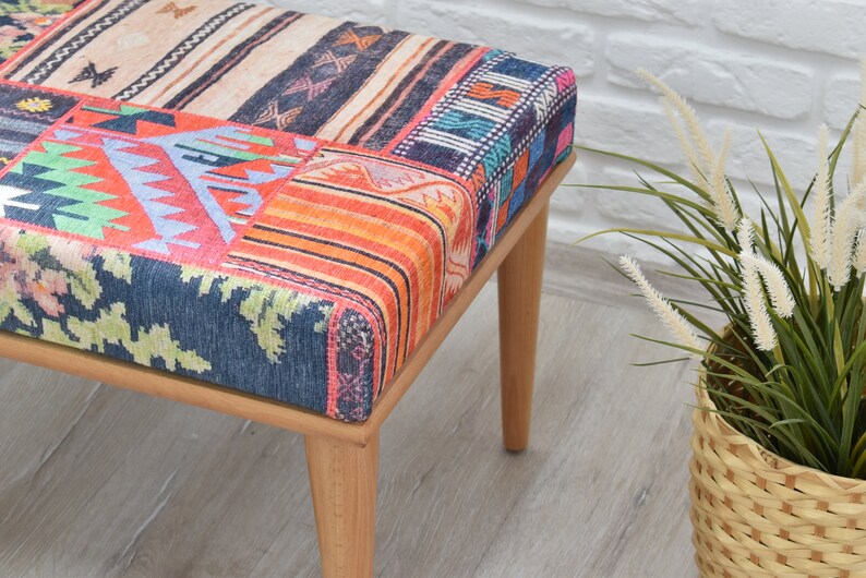 Pode incluir: Banqueta de madeira com assento em patchwork. O assento apresenta v&aacute;rios padr&otilde;es de tecido coloridos, incluindo motivos geom&eacute;tricos e florais. A banqueta tem quatro pernas de madeira clara e est&aacute; perto de uma planta decorativa.