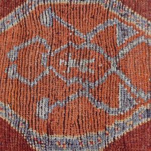 May include: Close-up of a textured, hand-woven rug with a geometric diamond pattern. The rug features a central orange diamond shape with a detailed gray and orange design, set against a deep red background. The rug's texture is visible.