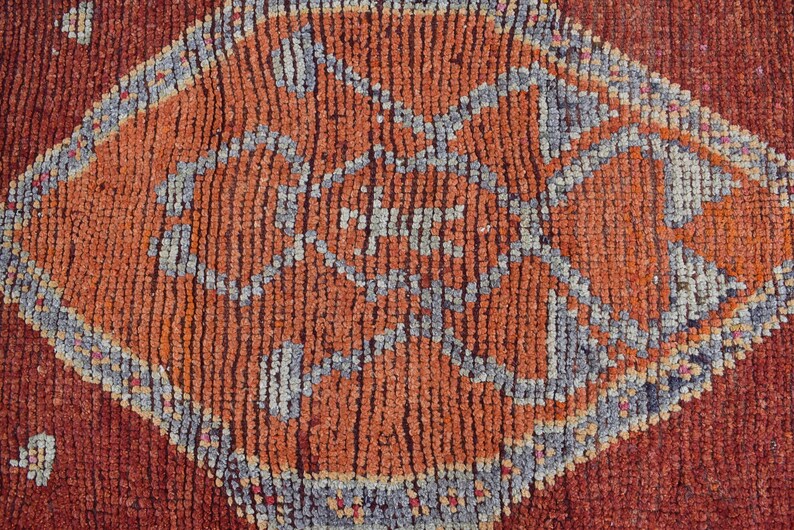 May include: Close-up of a textured, hand-woven rug with a geometric diamond pattern. The rug features a central orange diamond shape with a detailed gray and orange design, set against a deep red background. The rug's texture is visible.
