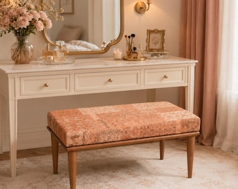May include: A rectangular bench with a cushioned seat featuring a peach and tan patterned fabric. The bench has four tapered wooden legs in a medium brown finish. It is positioned in front of a white vanity table with three drawers.