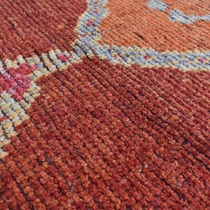 May include: Close-up of a hand-woven rug with a textured surface. The rug features a pattern of geometric shapes in shades of red, orange, pink, and beige, with a gray border.