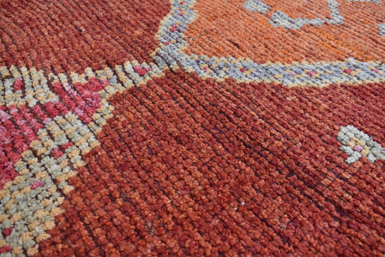 May include: Close-up of a hand-woven rug with a textured surface. The rug features a pattern of geometric shapes in shades of red, orange, pink, and beige, with a gray border.