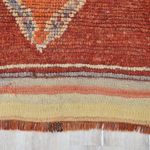 May include: Close-up of a handmade rug with a geometric design. The rug features a textured, reddish-orange field with a diamond pattern in orange, gray, and white. The bottom has horizontal stripes in beige, orange, and gray.