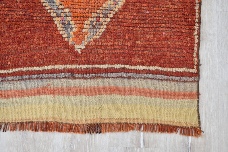 May include: Close-up of a handmade rug with a geometric design. The rug features a textured, reddish-orange field with a diamond pattern in orange, gray, and white. The bottom has horizontal stripes in beige, orange, and gray.