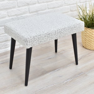 May include: A white fluffy ottoman with black legs. The ottoman is sitting on a light wood floor.