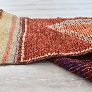 May include: A rolled-up, textured rug with a geometric design. The rug features a color palette of orange, red, beige, and purple. The rug has a fringed edge and is made of a woven material.