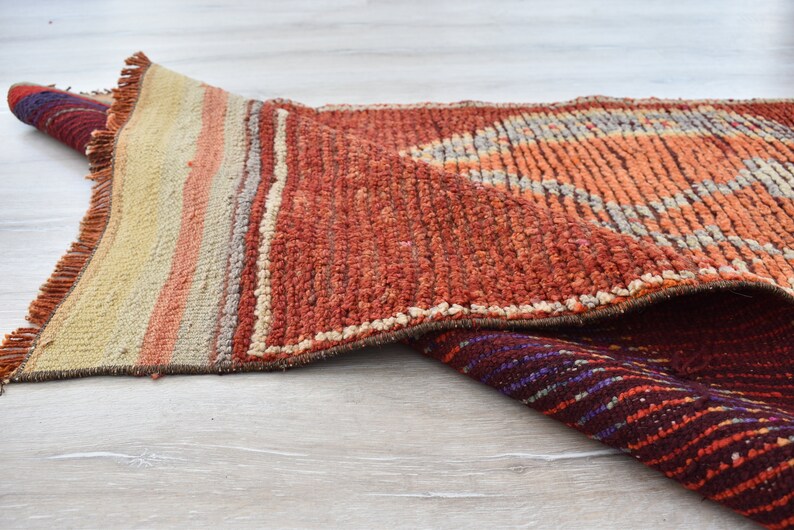 May include: A rolled-up, textured rug with a geometric design. The rug features a color palette of orange, red, beige, and purple. The rug has a fringed edge and is made of a woven material.