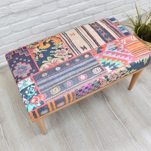 Pode incluir: Banqueta retangular com design de patchwork. O assento apresenta uma variedade de padr&otilde;es de tecido coloridos, incluindo desenhos florais, geom&eacute;tricos e listrados. A banqueta tem quatro pernas de madeira e est&aacute; colocada em um piso de madeira clara.