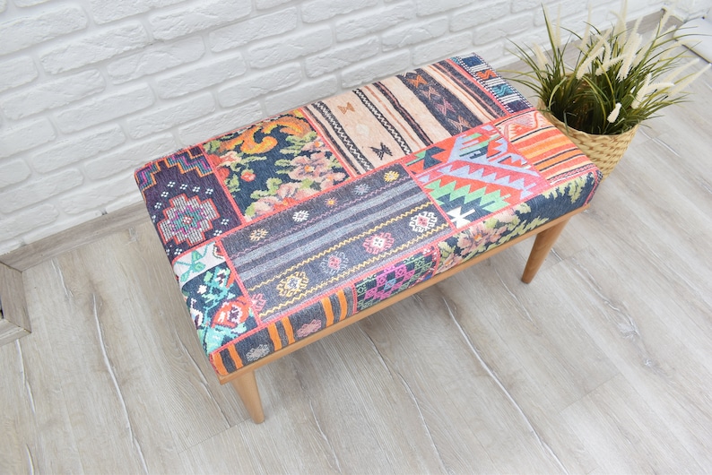 Pode incluir: Banqueta retangular com design de patchwork. O assento apresenta uma variedade de padr&otilde;es de tecido coloridos, incluindo desenhos florais, geom&eacute;tricos e listrados. A banqueta tem quatro pernas de madeira e est&aacute; colocada em um piso de madeira clara.