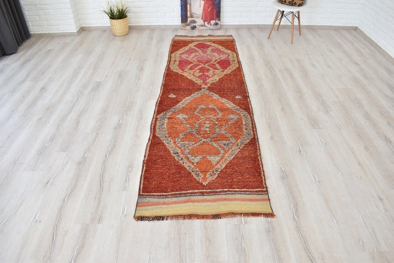 May include: A long, rectangular, hand-woven rug with a geometric pattern. The rug features a warm color palette of red, orange, and beige. The design includes diamond shapes and intricate details, suitable for a hallway or entryway.