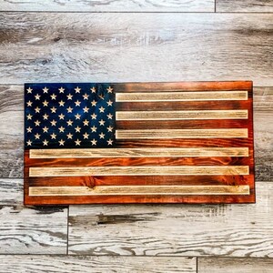 Könnte beinhalten: Eine amerikanische Holzflagge mit einer abgenutzten Oberfläche. Die Flagge ist in Rot, Weiß und Blau mit einem dunkelblauen Sternenfeld bemalt. Die Flagge hängt an einer hellbraunen Holzwand.
