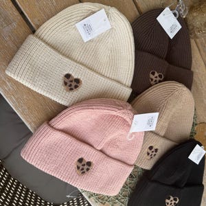 Könnte beinhalten: Eine Auswahl an gerippten Strickmützen in Creme, Braun, Rosa, Beige und Schwarz. Jede Mütze hat einen herzförmigen Aufnäher mit Leopardenmuster. Die Mützen liegen auf einer Holzoberfläche.