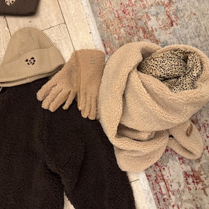 May include: A collection of winter accessories including a brown beanie, gloves, and a scarf. The beanie and gloves are tan, and the scarf is beige with a leopard print lining. A dark brown, button-up jacket is also included.