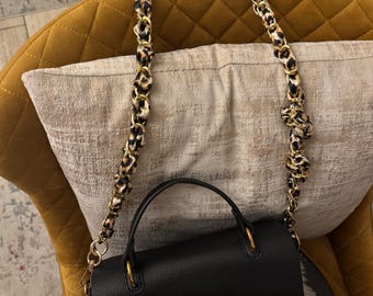 Puede incluir: Un bolso de mano de cuero negro con una correa de cadena dorada y un cierre en forma de H dorado. La correa tiene un estampado de leopardo.