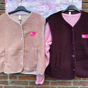Könnte beinhalten: Zwei ärmellose Westen aus strukturiertem Fleece-Material. Die linke Weste ist hellrosa, die rechte dunkelburgunderrot. Beide haben einen V-Ausschnitt, eine Knopfleiste mit vier Knöpfen auf der Vorderseite und ein kleines rechteckiges Etikett.