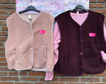 Könnte beinhalten: Zwei ärmellose Westen aus strukturiertem Fleece-Material. Die linke Weste ist hellrosa, die rechte dunkelburgunderrot. Beide haben einen V-Ausschnitt, eine Knopfleiste mit vier Knöpfen auf der Vorderseite und ein kleines rechteckiges Etikett.