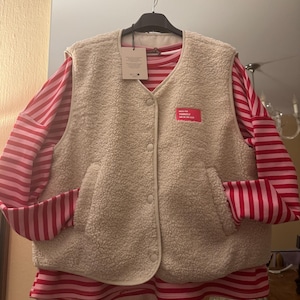 Könnte beinhalten: Eine beige Fleeceweste mit Knopfleiste und zwei Taschen, über einem langärmeligen rot-rosa gestreiften Hemd. Ein kleiner rosa rechteckiger Aufnäher mit weißem Text befindet sich auf der Weste. Das Outfit hängt an einem schwarzen Kleiderbügel.