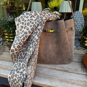 Puede incluir: Un bolso tipo cubo color topo con acabado texturizado de imitación cocodrilo, que presenta un cierre giratorio de metal dorado en una correa horizontal. Una bufanda larga y rectangular con estampado de leopardo gris y beige cuelga del asa.