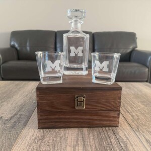 May include: A clear glass decanter with a stopper and two matching glasses, each etched with a white "M". The set sits on a wooden box with a metal clasp. The decanter set is a great gift for any occasion.