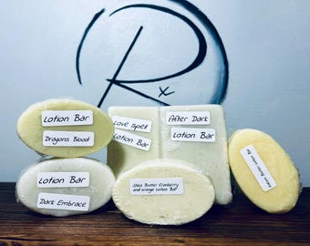 May include: Six different lotion bars with various scents, including Dragons Blood, Dark Embrace, Love Spell, After Dark, Shea Butter Cranberry and Orange, and Kokum Butter. The lotion bars are wrapped in clear plastic and are arranged on a wooden surface.