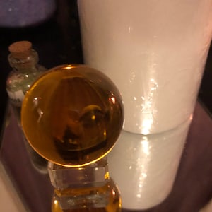 May include: A golden glass sphere sits on a clear glass stand. The sphere is reflecting light and appears to be filled with a liquid.