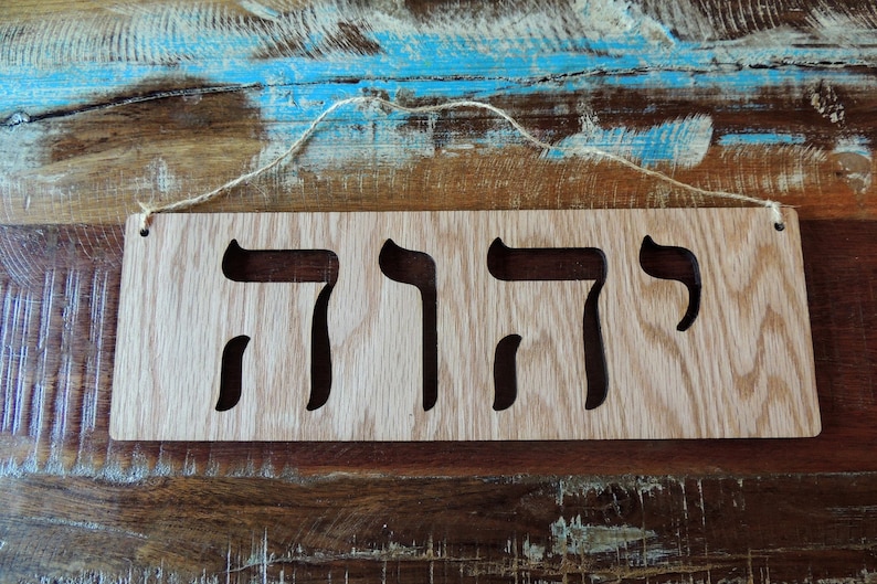 A stained rectangle Oak sign is shown on a table. In the center, the Hebrew words for YHWH are cut out. Holes are cut from the two top corners with twine strung across.