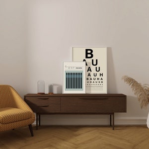 May include: A living room interior with a yellow armchair, a wooden console table with two framed prints, a glass cloche, and a vase with dried pampas grass. The print on the right has the text "BAUHAUS" repeated in black.