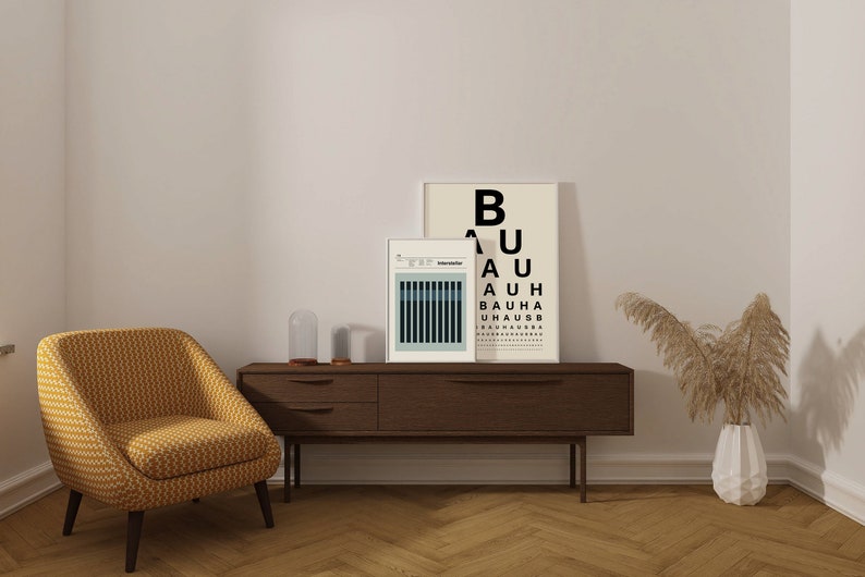 May include: A living room interior with a yellow armchair, a wooden console table with two framed prints, a glass cloche, and a vase with dried pampas grass. The print on the right has the text "BAUHAUS" repeated in black.