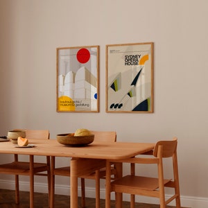 May include: Two framed posters hanging on a light beige wall above a wooden dining table with four chairs. The poster on the left features a white building with a red circle and blue circle. The text on the poster reads "bauhaus-archiv/ museum fur gestaltung". The poster on the right features a white building with a yellow circle and blue and green geometric shapes. The text on the poster reads "SYDNEY OPERA HOUSE".