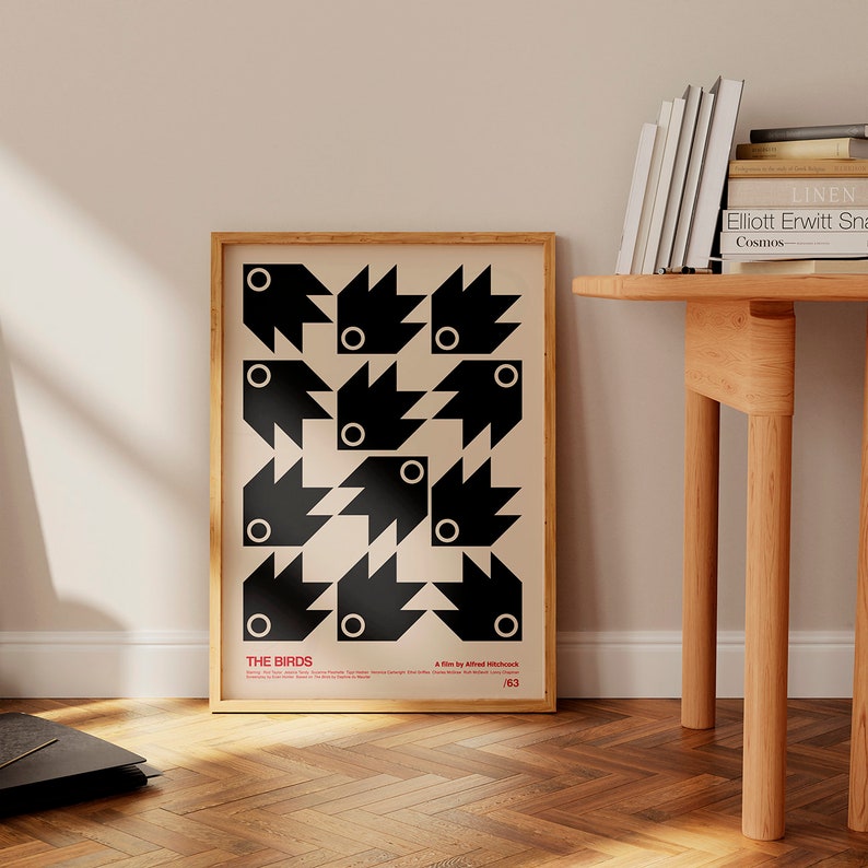 May include: A black and white poster featuring a geometric design of birds. The poster is framed in wood and has the title "THE BIRDS" and the text "A film by Alfred Hitchcock" at the bottom. The poster is hanging on a wall in a room with a wooden floor.