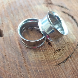 May include: Two silver metal ear gauges, one slightly larger than the other, are lying on a wooden surface.