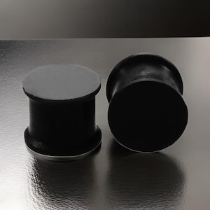 May include: Two black rubber ear gauges, one on top of the other, on a silver surface.