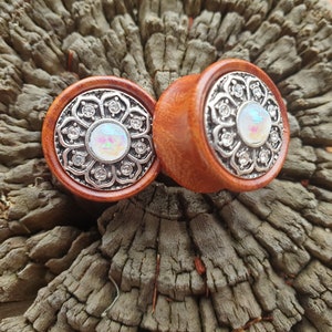 Puede incluir: Par de dilatadores de oreja de madera con una incrustación de metal plateado que presenta un diseño floral con una piedra blanca iridiscente en el centro.