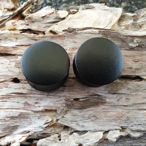 Può includere: Due tappi per orecchie in legno nero, noti anche come tappi, sono mostrati su un pezzo di legno invecchiato.