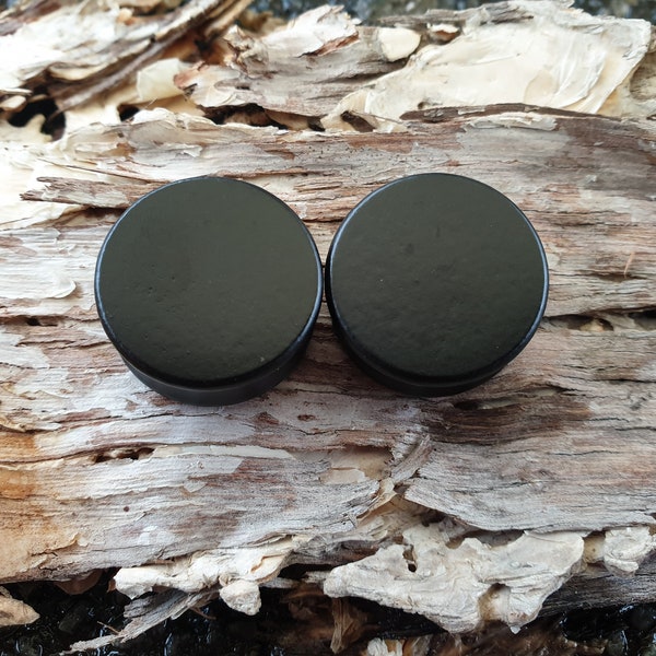 Könnte beinhalten: Zwei schwarze Holzohrstecker, auch bekannt als Ohrstöpsel, werden auf einem Stück Baumrinde gezeigt.