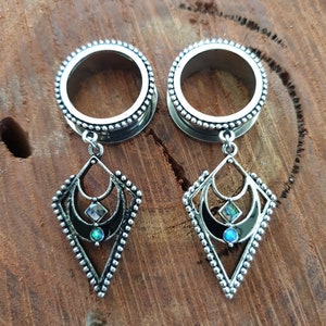 Puede incluir: Un par de dilatadores de oreja de plata con colgantes. Los colgantes son negros con detalles plateados y presentan un diseño de luna creciente con una ópalo y una pedrería transparente.