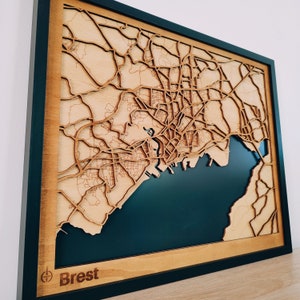 May include: A wooden map of Brest, France, with a dark blue background representing the water. The map is framed in black and has a wooden border. The city name 'Brest' is written in the lower left corner.
