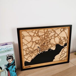 May include: A wooden map of Brest, France, with a black background and the word 'Brest' in the bottom left corner. The map is framed in black and has a light wood border.