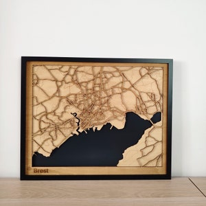 May include: A wooden map of Brest, France, with a black background and a black frame. The map is laser cut and shows the city's streets and waterways. The word 'Brest' is printed in the lower left corner of the map.