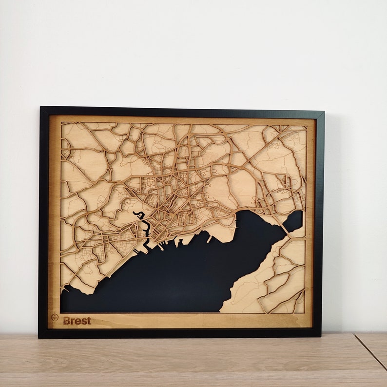May include: A wooden map of Brest, France, with a black background and a black frame. The map is laser cut and shows the city's streets and waterways. The word 'Brest' is printed in the lower left corner of the map.