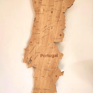May include: A wooden map of Portugal, showcasing the country's outline and internal regions. The map is made of light-colored wood, with the country's name and regions labeled in a dark brown font. The map includes cities and landmarks.