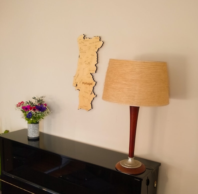 May include: A wooden map of Portugal hangs on a white wall, with city names etched into the wood. A tan lampshade sits atop a dark wood and silver lamp base. A bouquet of flowers sits in a gray vase on a black piano.