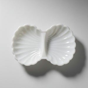 May include: A white, divided dish with a shell-like design. The dish is made of a glossy material and has a scalloped edge. The two sections are separated by a raised divider. The dish is empty and sits on a white surface.