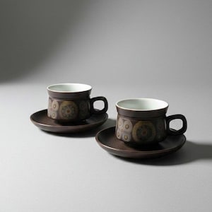 May include: Two brown ceramic teacups with matching saucers. The cups have a dark brown exterior with a floral pattern and white interiors. The saucers are a solid brown color. The cups have a handle.