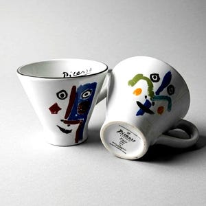 May include: Two white ceramic mugs with abstract Picasso-style designs. One mug features a face with blue, red, and black accents, and the word "Picasso" is written on the rim. The other mug has colorful abstract shapes.
