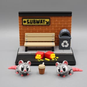 May include: A miniature diorama featuring a subway scene with a brick wall, bench, and trash can. Two gray rats with pink tails flank a coffee cup. Scattered french fries and a sign that says "SUBWAY" complete the scene.