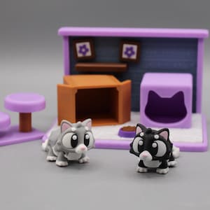 Puede incluir: Un juego de miniaturas con temática de gatos con dos gatos de juguete, una casa para gatos morada y una caja para gatos marrón. Los gatos son grises y negros con ojos grandes. El juego incluye una cama para gatos morada y dos taburetes morados.