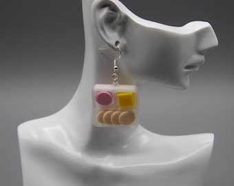 May include: A pair of novelty earrings shaped like a clear plastic container, each filled with miniature food items. The earrings feature a pink circle, a yellow square, and several small, round, beige-colored items. The earrings hang from silver-colored hooks.